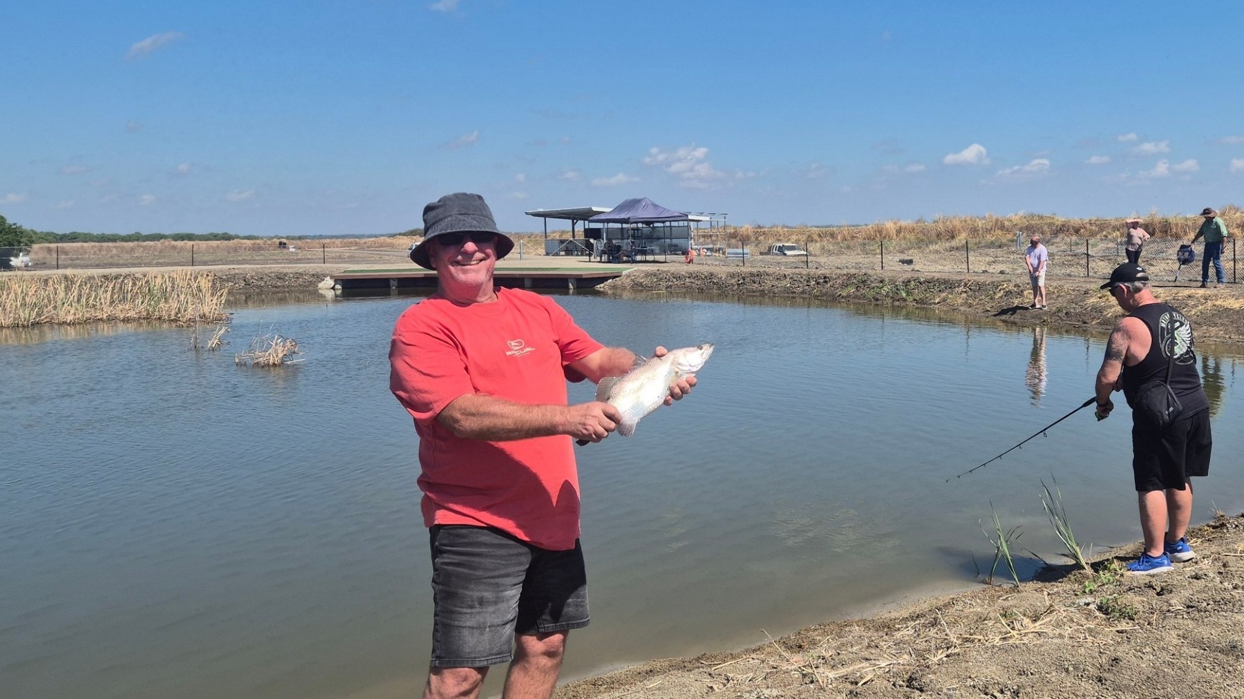 Man in red shirt holding fish at a pond; others fishing nearby under clear sky.