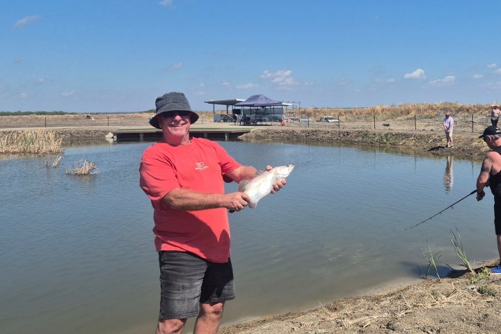 Man in red shirt holding fish at a pond; others fishing nearby under clear sky.