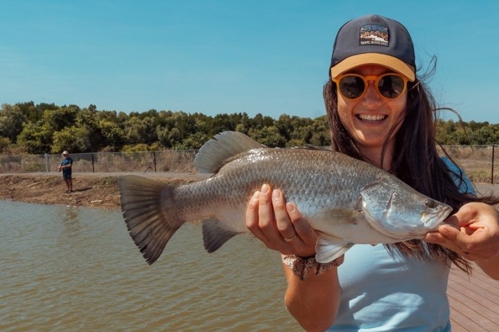 Woman in cap holding a fish, smiling, with trees in the background.