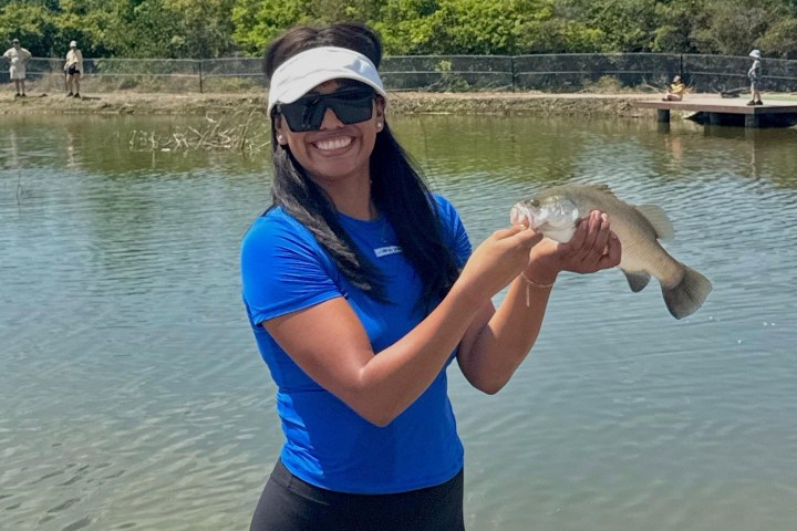 Smiling person in blue shirt holding a fish by the water.