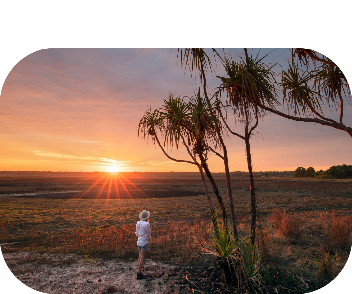 Person in white clothes and hat watching a sunset over a field with a tree.