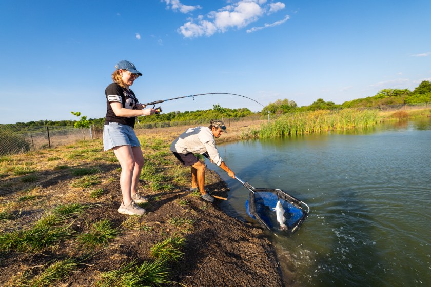 Two people fishing at a lakeside, one holding a rod, the other nets a fish.